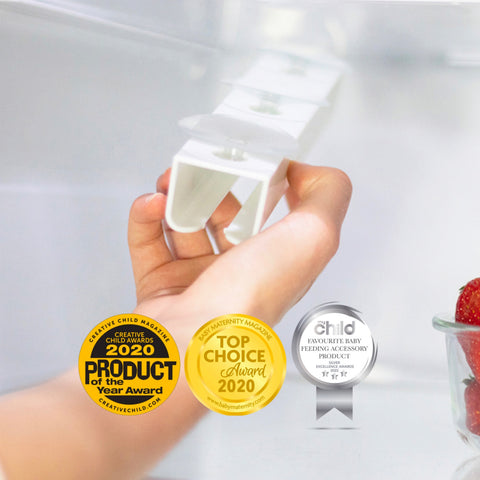 A hand holds a white plastic baby bottle clip against a soft white background. Three award badges are overlaid at the bottom left: a gold Creative Child Wearable Product of the Year Award, a gold Best University Magazine Top Choice Award, and a silver Child Favorite Baby Feeding Accessory Product award.