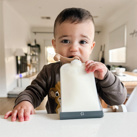 A baby wearing a brown long-sleeve shirt with a cartoon bear graphic sits at a white table, holding a white feeding pouch with a wooden spoon and drinking from a small cup. The background shows a softly blurred modern home interior.